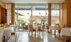 Ett modernt rum med dukade bord och stolar, samt stora fönster som vätter mot en uteplats med träd och möbler. (AI-genererad text)