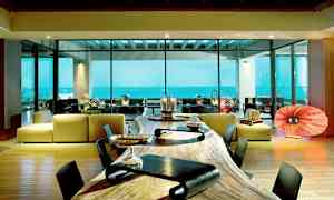 Ett modernt rum med stora glasfönster som visar utsikt över havet. Det finns en lång träbord i förgrunden och en sittgrupp med en modern design i bakgrunden. (AI-genererad text)