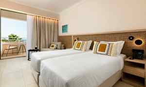 Ett hotellrum med två enkelsängar, en sittgrupp och en balkong som visar utsikt över grönska och himmel. (AI-genererad text)