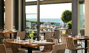 Utsikt över en modern restaurang med rottingstolar och bord, med en trädgård och havet i bakgrunden. (AI-genererad text)