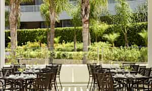 Flera bord med stolar i en utomhusmiljö omgiven av gröna växter och palmer. (AI-genererad text)