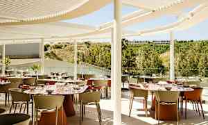 En utomhusrestaurang med matbord och stolar under en modern vit solskärm. Bakgrunden visar grönska och berg. (AI-genererad text)