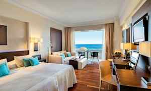 Hotellrum med dubbelsäng, sittgrupp och utsikt mot havet. (AI-genererad text)