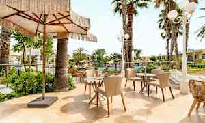 En utomhusterrass med bord och stolar omgiven av palmer och grönska. En stor solparasoll ger skugga. (AI-genererad text)