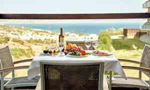 Ett bord med duk, glas och en flaska vin, omgivet av stolar, med utsikt över ett landskap och hav. (AI-genererad text)