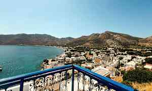 Vy från en balkong med blått räcke över en kuststad med vita byggnader och omgivande berg. (AI-genererad text)