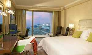 Hotellrum med en dubbelbädd säng, stolar och utsikt över balkonger och havet genom stora fönster. (AI-genererad text)