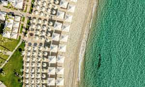 Flygbild av en strand med solparasoller i rader och klart turkost vatten. (AI-genererad text)
