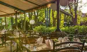 Uteservering med dukade bord och stolar under ett tak med gröna träd i bakgrunden. (AI-genererad text)