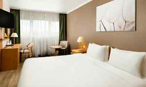 Ett hotellrum med en stor dubbelsäng, skrivbord, stol, fåtölj och fönster med ljusa gardiner. (AI-genererad text)