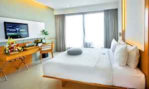 En modern hotellrum med en stor bekväm säng, skrivbord med blomarrangemang och en TV på väggen. Fönster med ljusa gardiner släpper in dagsljus. (AI-genererad text)