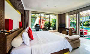 Ett hotellrum med en stor säng med vita sängkläder och röda kuddar framför ett stort fönster som vetter mot en trädgård. (AI-genererad text)