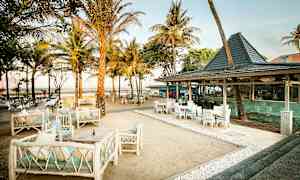 En uteservering vid en sandstrand med bord, stolar och soffor omgiven av palmer. (AI-genererad text)