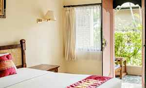 Ett ljust hotellrum med en säng täckt av vitt sänglinne och en dekorativ röd löpare. Ett fönster med gardiner och en öppen dörr leder till en uteplats med grönska. (AI-genererad text)