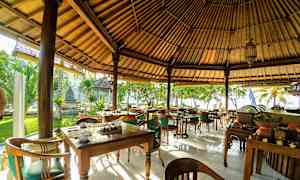 En utomhusrestaurang med träbord och stolar under ett halmtak, omgiven av grönska och belägen nära stranden. (AI-genererad text)