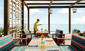 Restaurang vid havet med träbord och stolar som har randiga dynor. (AI-genererad text)