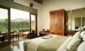 Ett sovrum med en säng som är bäddad med grön och vit sängkläder. Rummet har en garderob i trä, nattygsbord och en stor spegel. Glasdörrar leder till en balkong med utsikt över grönskande natur. (AI-genererad text)