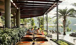 En utomhusrestaurang med träbord och stolar, tak av träplankor och gröna växter. Pool och palmer syns i bakgrunden. (AI-genererad text)