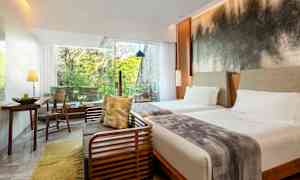 Ett hotellrum med två sängar, en sittgrupp, och stora fönster som släpper in ljus och ger utsikt över grönskande träd. (AI-genererad text)