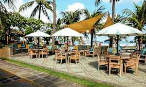 En uteservering nära en strand med träbord, stolar, parasoller och palmer. (AI-genererad text)