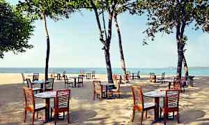 Utomhusbord och stolar i trä placerade på en sandstrand med havet i bakgrunden under träd. (AI-genererad text)