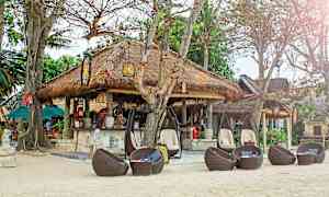 En strandrestaurang med halmtak, omgiven av träd och sittplatser i flätade rottingmöbler. (AI-genererad text)