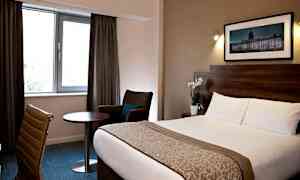 Hotellrum med säng, stol, bord och fönster med gardiner. (AI-genererad text)