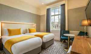 Två sängar med gula sängöverkast och kuddar i ett rum med bord, stolar och ett fönster. (AI-genererad text)