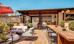 En utomhusterrass med sittplatser, bord och grönska under en klarblå himmel. (AI-genererad text)