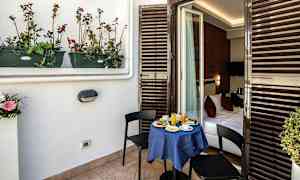 En balkong med ett litet bord dukat för frukost och två stolar. Bordet är täckt med en blå duk och det leder in till ett sovrum. (AI-genererad text)