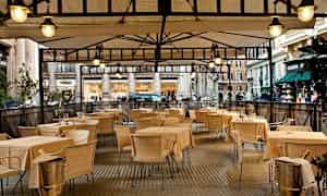 Bild av en uteservering med bord och stolar under ett täckt tak. (AI-genererad text)