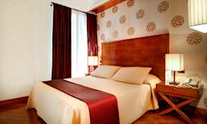 Sovrum med en stor säng täckt med beige och vinröda sängkläder, två sidobord med lampor och dekorerad tapet på väggen. (AI-genererad text)