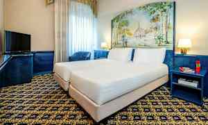 Ett hotellrum med två enkelsängar placerade intill varandra, ett mönstrat golv och ett målning som pryder väggen bakom sänggavlarna. Det finns en TV, en blå fåtölj och ett nattduksbord med böcker och pennor. (AI-genererad text)