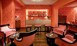 Bild som visar en elegant utrymme inrett som en cocktailbar med mörka möbler, rosa dynor och flaskor på en hylla. (AI-genererad text)