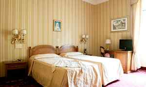 Ett hotellrum med två enkelsängar täckta med beige sängkläder, dekorerade väggar med tavlor, och möbler inklusive bord, stolar och en TV. (AI-genererad text)