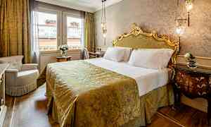 Ett elegant sovrum med en uppsatt säng med dekorerad gavel, täckt av ett gyllene överkast. Sidan av rummet har två fönster och ett bord med en blomvas. (AI-genererad text)