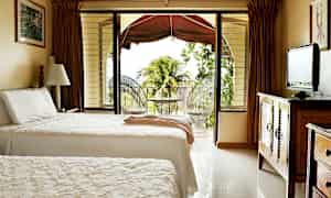 Ett hotellrum med två enkelsängar, en TV på en träbyrå och en öppning till en balkong med utsikt över växtlighet och palmträd. (AI-genererad text)