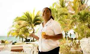 En person håller två glas, ett med vin och ett med juice, vid ett bord utomhus nära stranden och omgiven av palmer. (AI-genererad text)