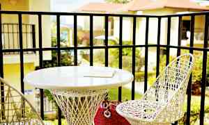 En balkong med två vita flätade stolar och ett runt bord. På bordet ligger en bok. Utsikt över grönska och byggnader i bakgrunden. (AI-genererad text)