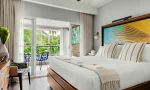 Hotellrum med en vit bädd, dekorativ väggkonst med palmblad, utsikt mot en balkong med grönskande miljö. (AI-genererad text)