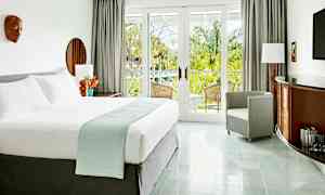 Ett ljust och elegant hotellrum med en stor säng, fåtölj, och möbler med trädetaljer. Rummet har stora glasdörrar som leder till en balkong med utsikt över grönskande palmer. (AI-genererad text)