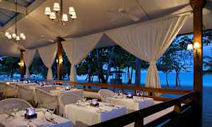 En restaurang med bord dukade med vita dukar vid stranden under ett kvällsbelyst tält. (AI-genererad text)