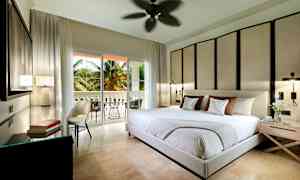 Ett modernt sovrum med stor säng, dekorativ väggpanel och öppning till en balkong med palmer utanför. (AI-genererad text)