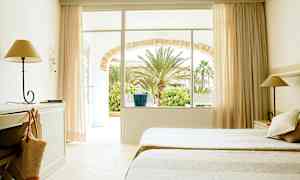 Ett hotellrum med två sängar, skrivbord och lampa. Fönster och dörr leder till en trädgård med palmer. (AI-genererad text)