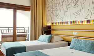 Ett hotellrum som visar två enkelsängar med turkosa dekorationer, bakom sängarna finns en vägg med konstnärlig dekoration och en liten lampett. (AI-genererad text)