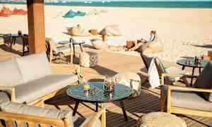 Uteplats med sittmöbler och bord, i bakgrunden en sandstrand och hav. (AI-genererad text)