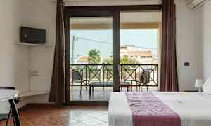 Ett rum med en säng, en liten TV på väggen och en balkong med utsikt över byggnader och palmer. (AI-genererad text)