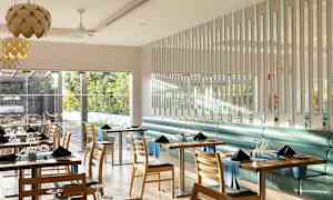 Ett modernt och ljusrestauranginteriör med bord och stolar, där fönster vetter mot ett grönskande utomhusområde. (AI-genererad text)