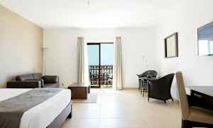 Ett hotellrum med ljus inredning. En säng, soffa, bord och stolar. En balkong med utsikt visas genom glasdörrar. (AI-genererad text)