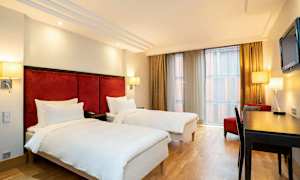 Ett hotellrum med två enkelsängar, skrivbord, stol och ett stort fönster med gardiner. (AI-genererad text)